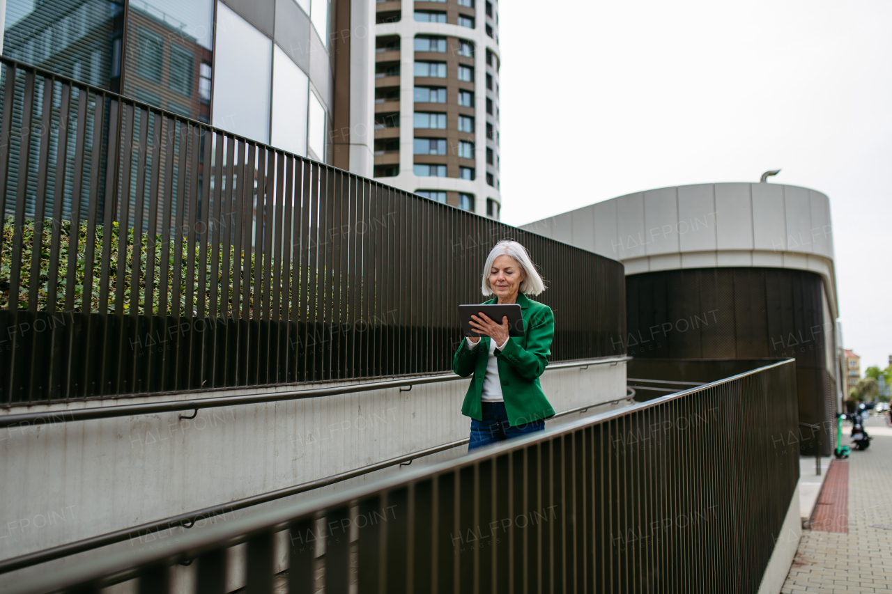 Mature businesswoman scrolling on tablet. Beautiful older woman with gray hair walking down the city street.