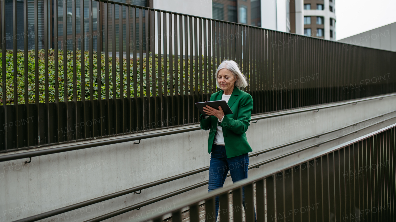 Mature businesswoman scrolling on tablet. Beautiful older woman with gray hair walking down the city street.