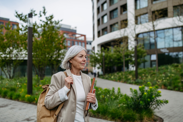 Mature businesswoman scrolling on smartphone, going home from work. Beautiful older woman with gray hair walking down the city street.