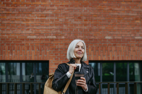 Portrait of stylish mature woman with gray hair on city street, outdoor. Older woman in leather jacket with soft smile on face.