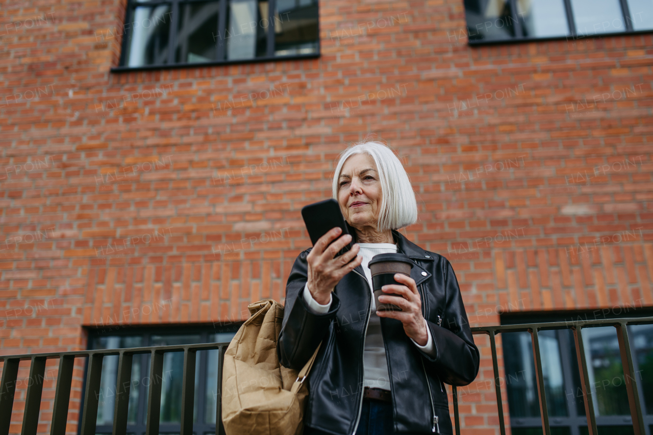 Portrait of stylish mature woman with gray hair on city street, , holding smartphone and travel mug with coffee, tea. Older woman in leather jacket with soft smile on face.