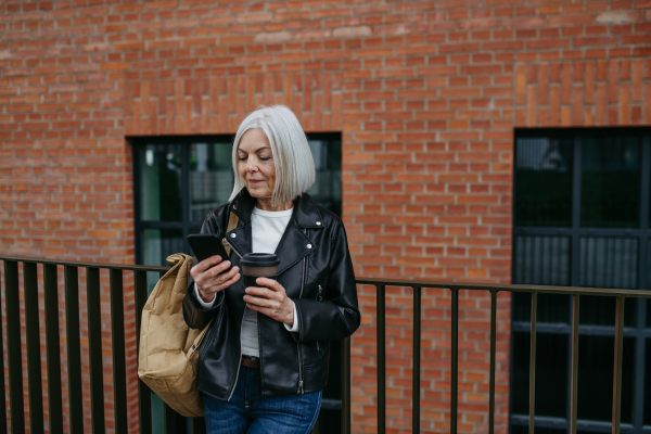 Portrait of stylish mature woman with gray hair on city street, outdoor. Older woman in leather jacket with soft smile on face.