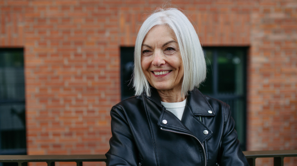 Portrait of stylish mature woman with gray hair on city street, outdoor. Older woman in leather jacket with soft smile on face.