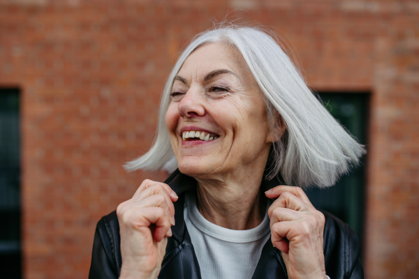 Portrait of stylish mature woman with gray hair on city street, outdoor. Older woman in leather jacket smiling.