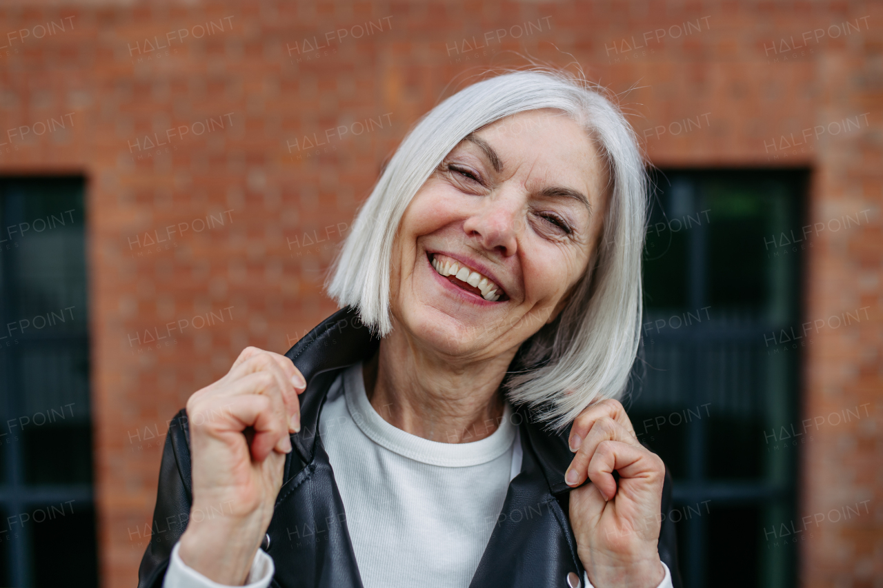 Portrait of stylish mature woman with gray hair on city street, outdoor. Older woman in leather jacket with soft smile on face.