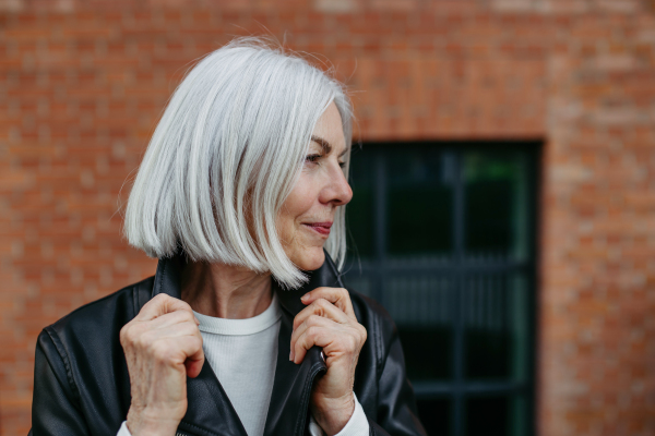 Portrait of stylish mature woman with gray hair on city street, outdoor. Older woman in leather jacket with soft smile on face.