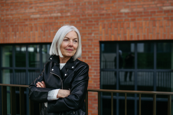 Portrait of stylish mature woman with gray hair on city street, outdoor. Older woman in leather jacket with soft smile on face.