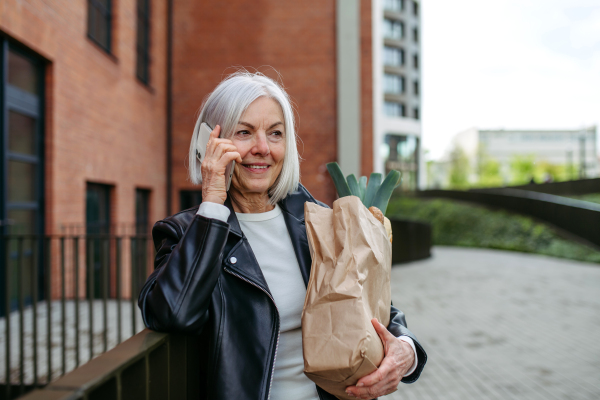 Mature woman making phone call, going home from supermaket with a gorceries. Beautiful older woman in leather jacket with gray hair standing on city street.