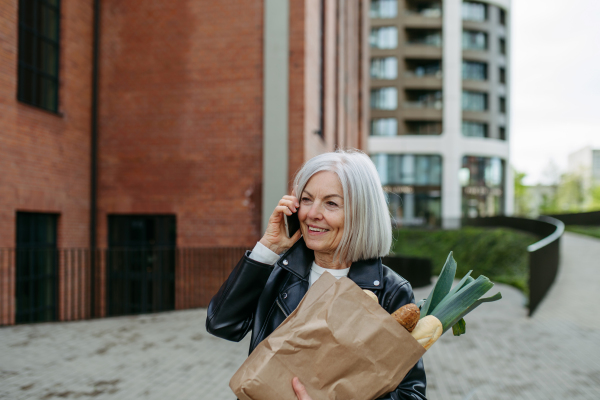 Mature woman making phone call, going home from supermaket with a gorceries. Beautiful older woman in leather jacket with gray hair standing on city street.