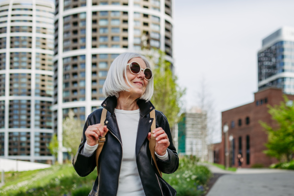 Portrait of stylish mature woman with gray hair on city street. Older woman in sunglasses waiting for public transport, urban commuter.