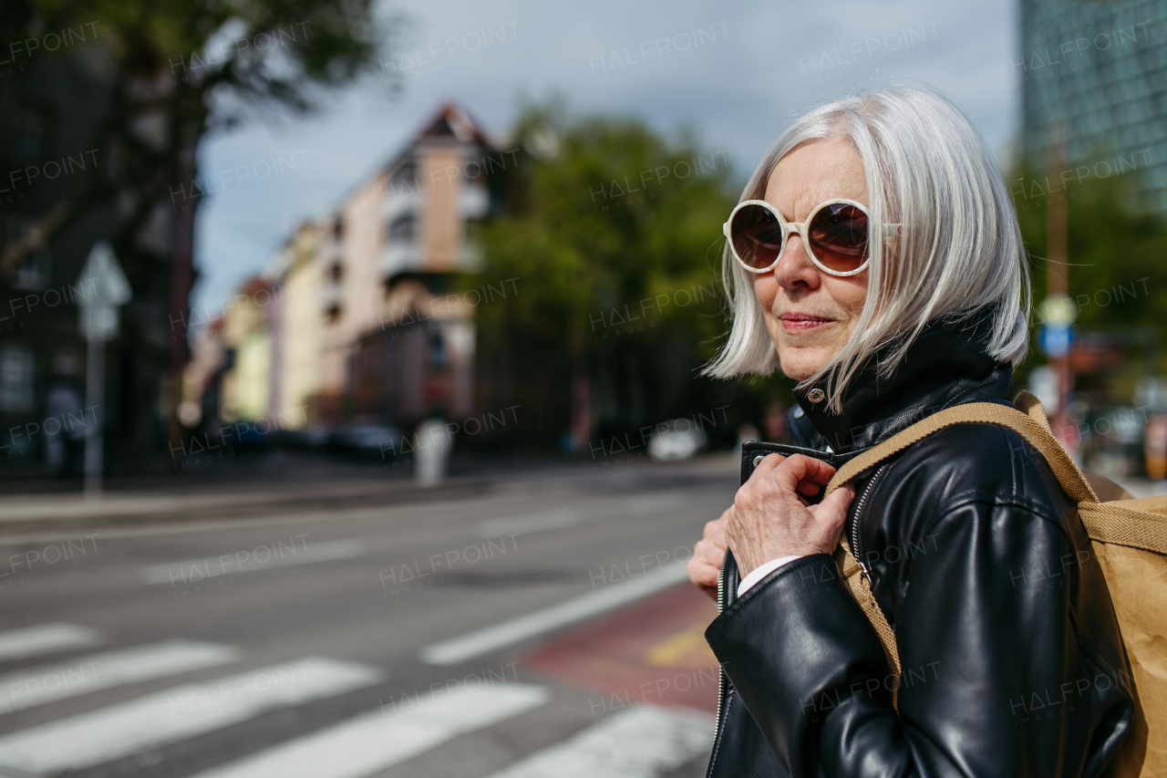 Portrait of stylish mature woman with gray hair on city street. Older woman in sunglasses waiting for public transport, urban commuter.