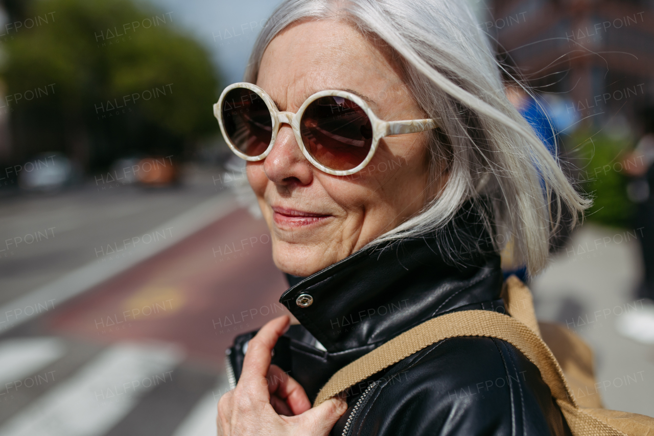 Portrait of stylish mature woman with gray hair on city street. Older woman in sunglasses waiting for public transport, urban commuter.