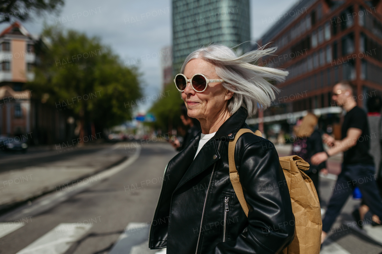 Portrait of stylish mature woman with gray hair on city street. Older woman in sunglasses, leather jacket crossing a road on pedestrian crossing.