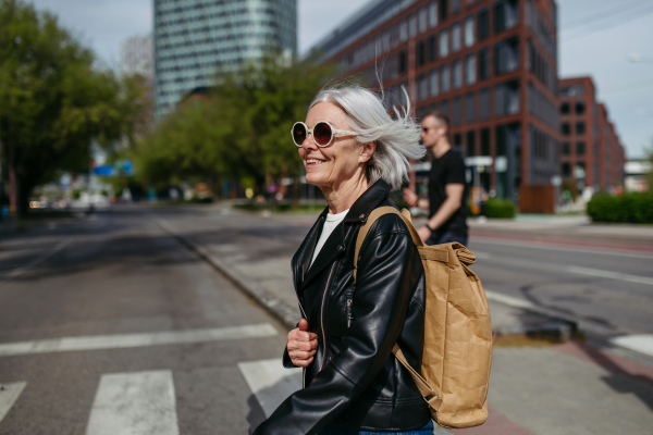 Portrait of stylish mature woman with gray hair on city street. Older woman in sunglasses waiting for public transport, urban commuter.