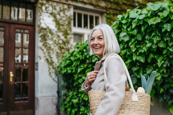 Stylish mature woman with gray hair going home from supermarket, standing in front her house with bag full of groceries.