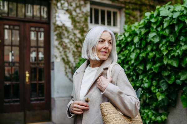 Stylish mature woman with gray hair going home from supermarket, standing in front her house with bag full of groceries.