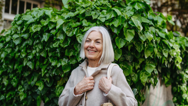 Stylish mature woman with gray hair going home from supermarket, standing in front her house with bag full of groceries.