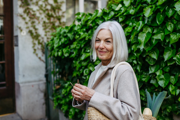 Stylish mature woman with gray hair going home from supermarket, standing in front her house with bag full of groceries.