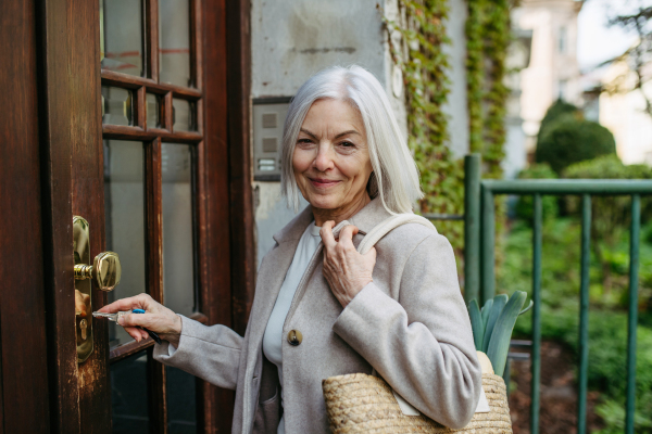 Stylish mature woman going home from supermarket, standing in front her house with bag full of groceries, unlocking door with key.