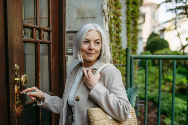 Stylish mature woman going home from supermarket, standing in front her house with bag full of groceries, unlocking door with key.