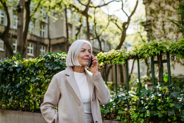 Portrait of stylish mature woman with gray hair on a city street, making phone call on smartphone.