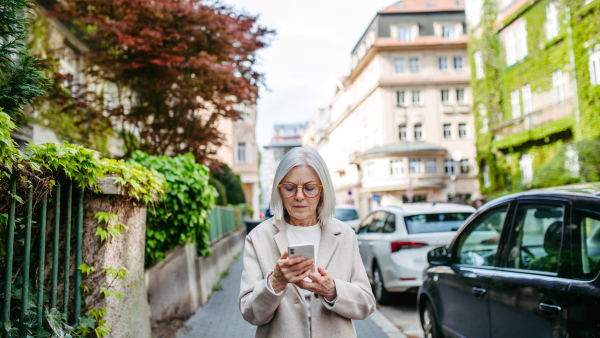 Mature woman holding smartphone, scrolling while walking on city street. Beautiful older woman with gray hair outdoors.