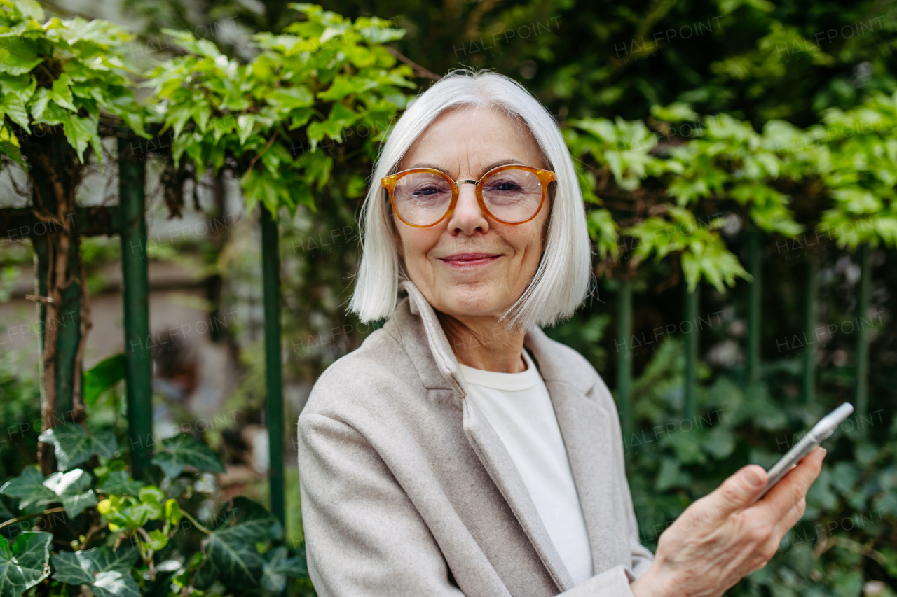 Mature woman holding smartphone, scrolling while relaxing in city park, sitting on bench.Beautiful older woman with gray hair outdoors.