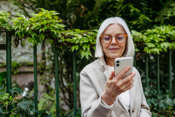 Mature woman holding smartphone, scrolling while relaxing in city park, sitting on bench.Beautiful older woman with gray hair outdoors.