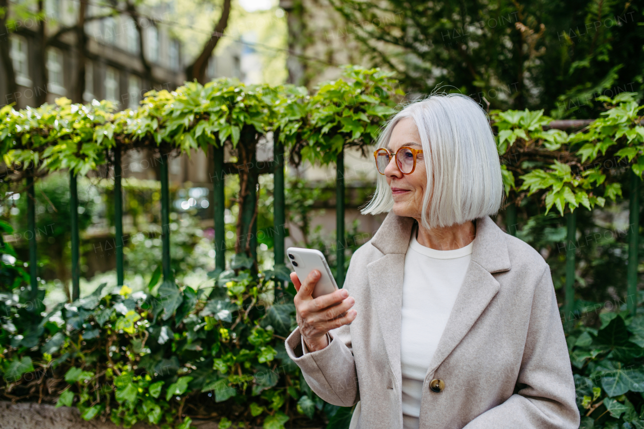 Mature woman holding smartphone, scrolling while relaxing in city park, sitting on bench.Beautiful older woman with gray hair outdoors.