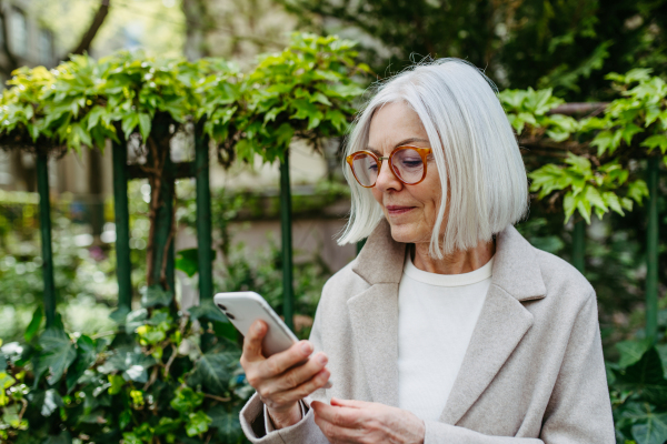 Mature woman holding smartphone, scrolling while relaxing in city park, sitting on bench.Beautiful older woman with gray hair outdoors.