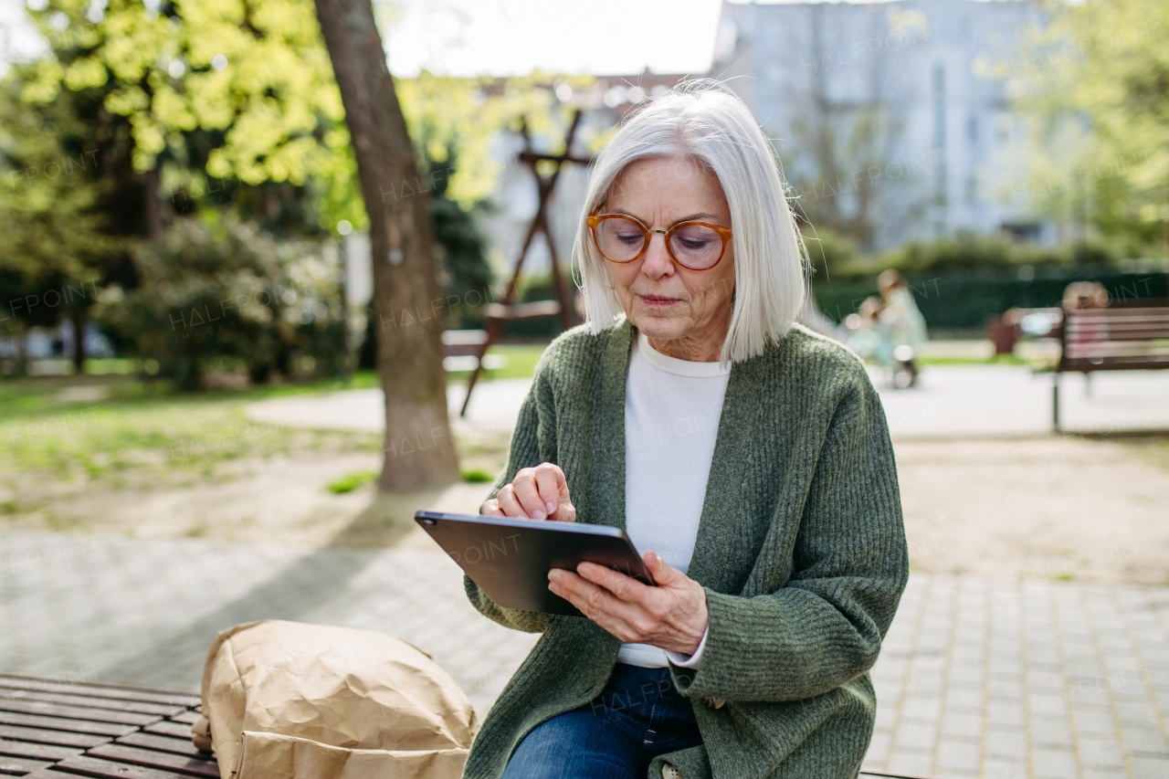 Mature woman sitting on park bench, working on tablet. Beautiful older woman with eyeglasses shopping online while outdoors.