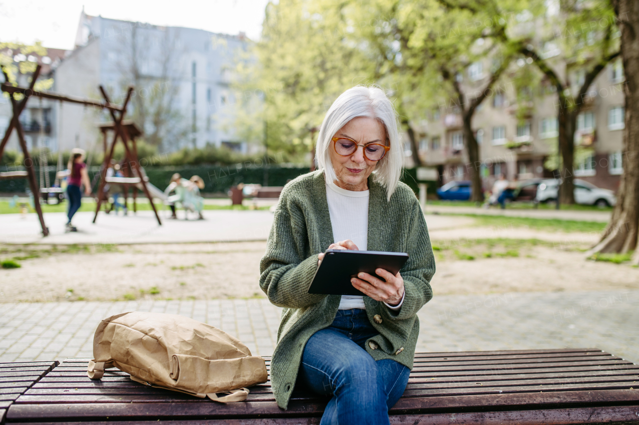 Mature woman sitting on park bench, working on tablet. Beautiful older woman with eyeglasses shopping online while outdoors.