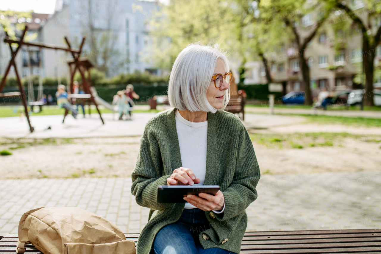 Mature woman sitting on park bench, working on tablet. Beautiful older woman with eyeglasses shopping online while outdoors.