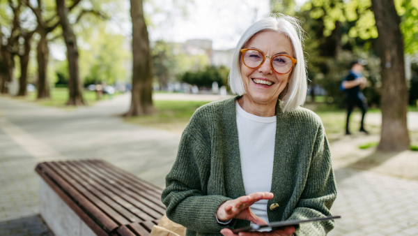 Mature woman sitting on park bench, working on tablet. Beautiful older woman with eyeglasses shopping online while outdoors.