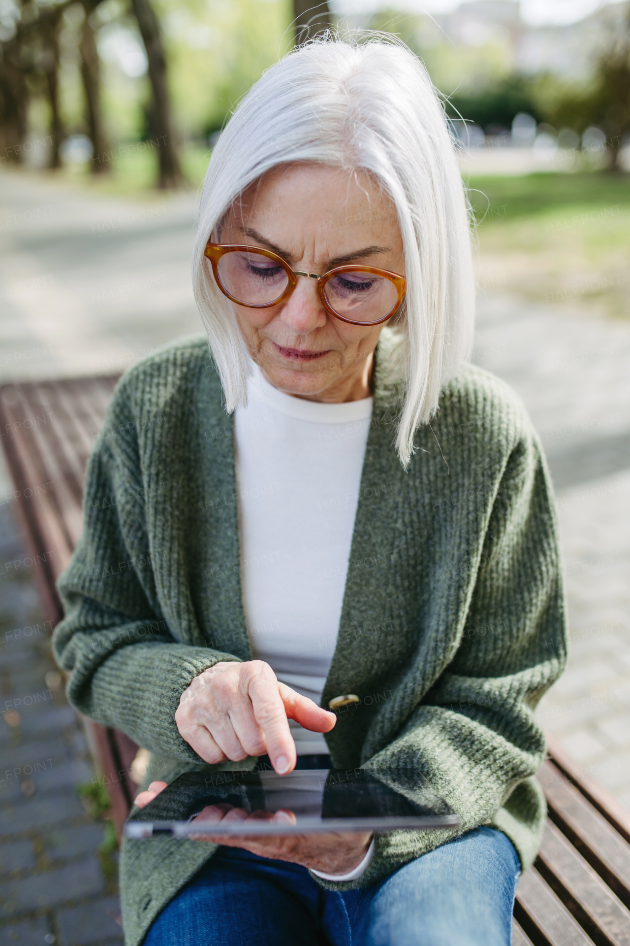 Mature woman sitting on park bench, working on tablet. Beautiful older woman with eyeglasses shopping online while outdoors.