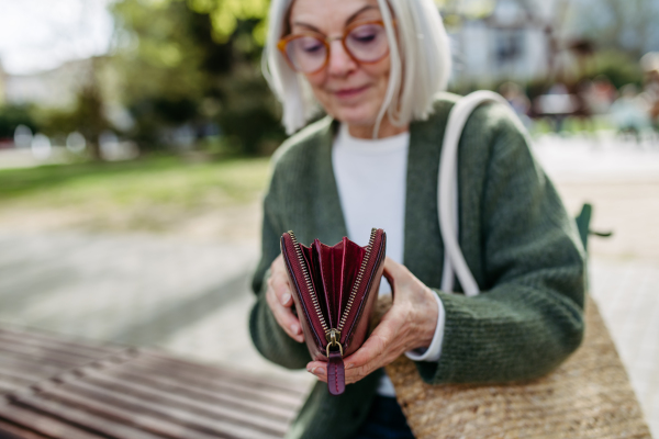 Mature woman with gray hair sitting on park bench, opening her wallet and looking inside.