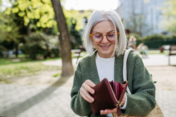 Mature woman with gray hair sitting on park bench, opening her wallet and looking inside.