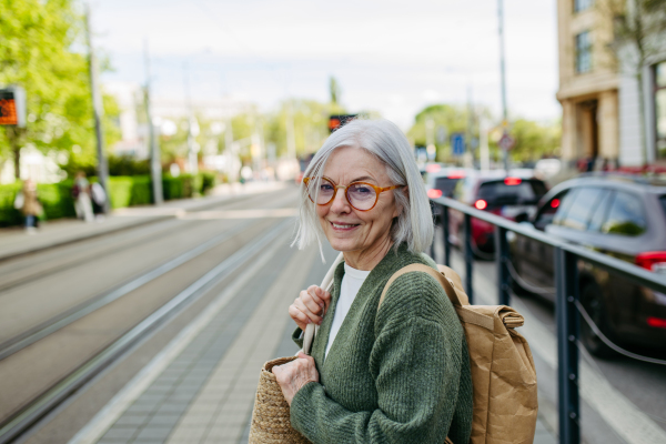 Portrait of stylish mature woman with gray hair on city street. Older woman with eyeglasses waiting for public transport.