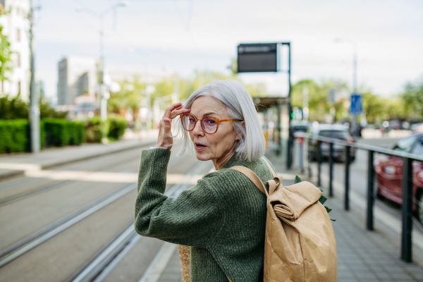 Portrait of stylish mature woman with gray hair on city street. Older woman with eyeglasses waiting for public transport.