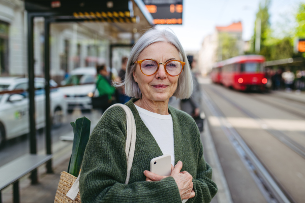 Portrait of stylish mature woman with gray hair on city street. Older woman with eyeglasses waiting for public transport.