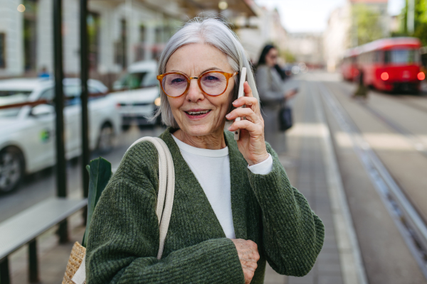 Portrait of stylish mature woman with gray hair on city street. Older woman making phone call while waiting for public transport, bus.