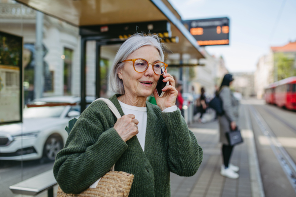 Portrait of stylish mature woman with gray hair on city street. Older woman making phone call while waiting for public transport, bus.