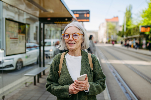 Portrait of stylish mature woman with gray hair on city street. Older woman with eyeglasses waiting for public transport, holding smartphne.