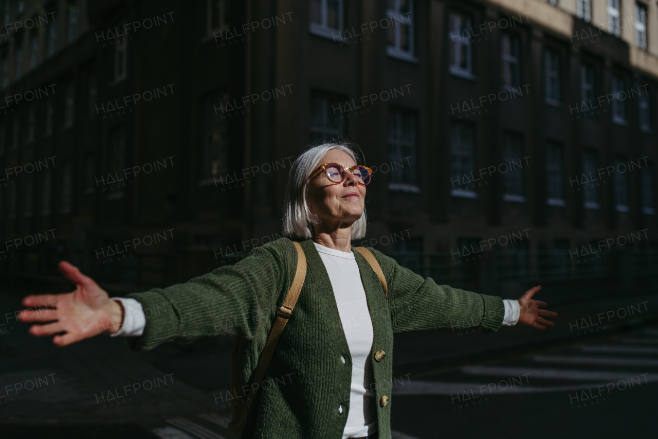 Portrait of beautiful mature woman standing on city street with open arms, enjoying warm sunshine on face. Woman with eyeglasses, gray hair and closed eyes is outdoors.