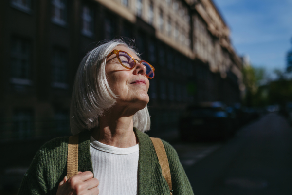 Portrait of beautiful mature woman standing on city street, enjoying warm sunshine on face. Woman with eyeglasses, gray hair and closed eyes is outdoors.