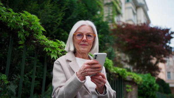 Mature woman holding smartphone, scrolling while relaxing in city park, sitting on bench. Beautiful older woman with gray hair outdoors.