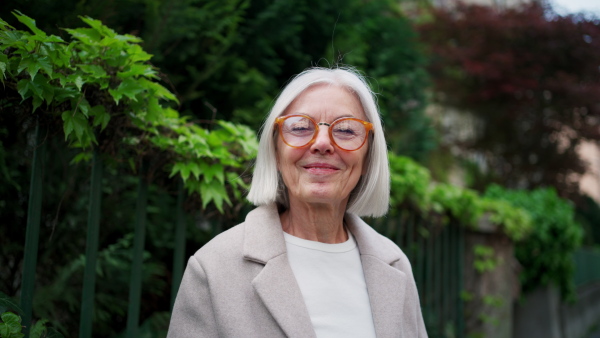 Mature woman holding smartphone, scrolling while relaxing in city park, sitting on bench. Beautiful older woman with gray hair outdoors.