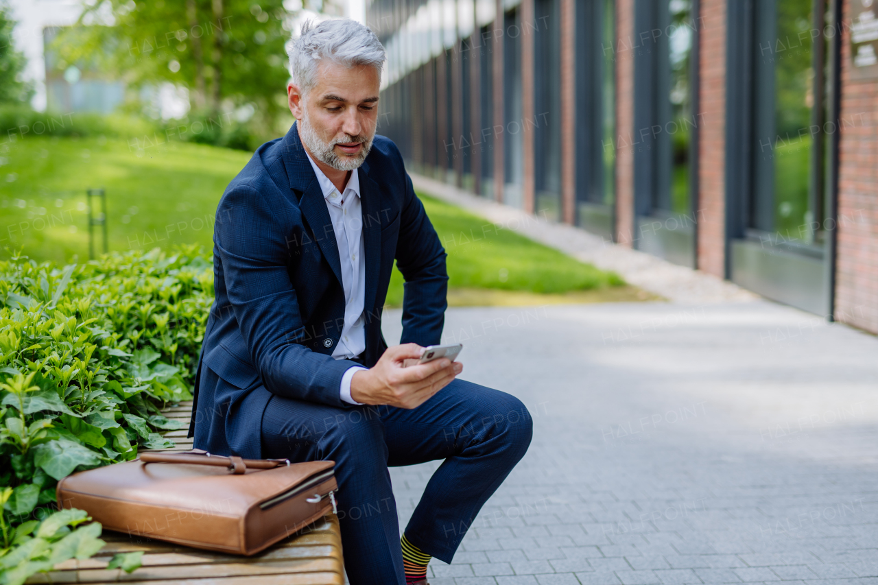 A successful mature businessman using smartphone while resting on street, sitting on bench and using mobile application.