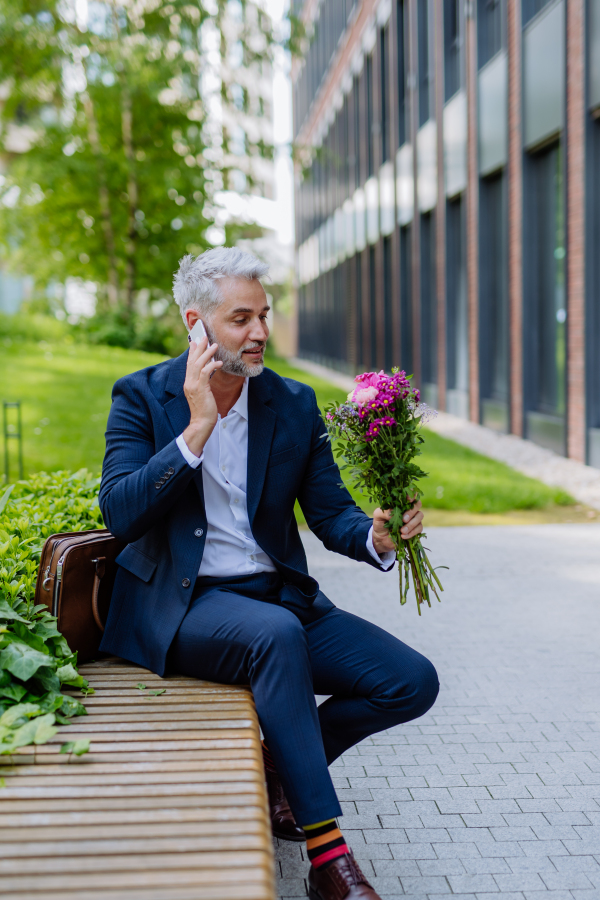 A portrait of happy mature businessman with flower bouquet sitting on bench and waiting for girlfriend.