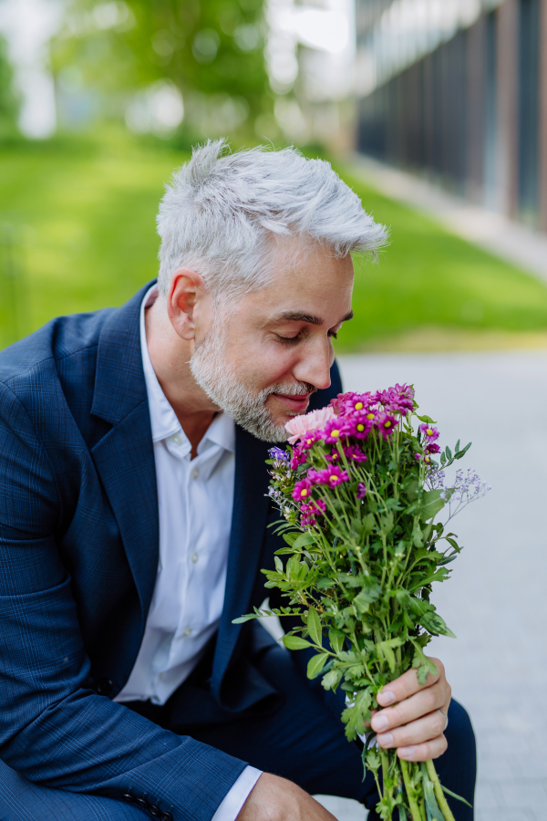 A portrait of happy mature businessman with flower bouquet sitting on bench and waiting for girlfriend.